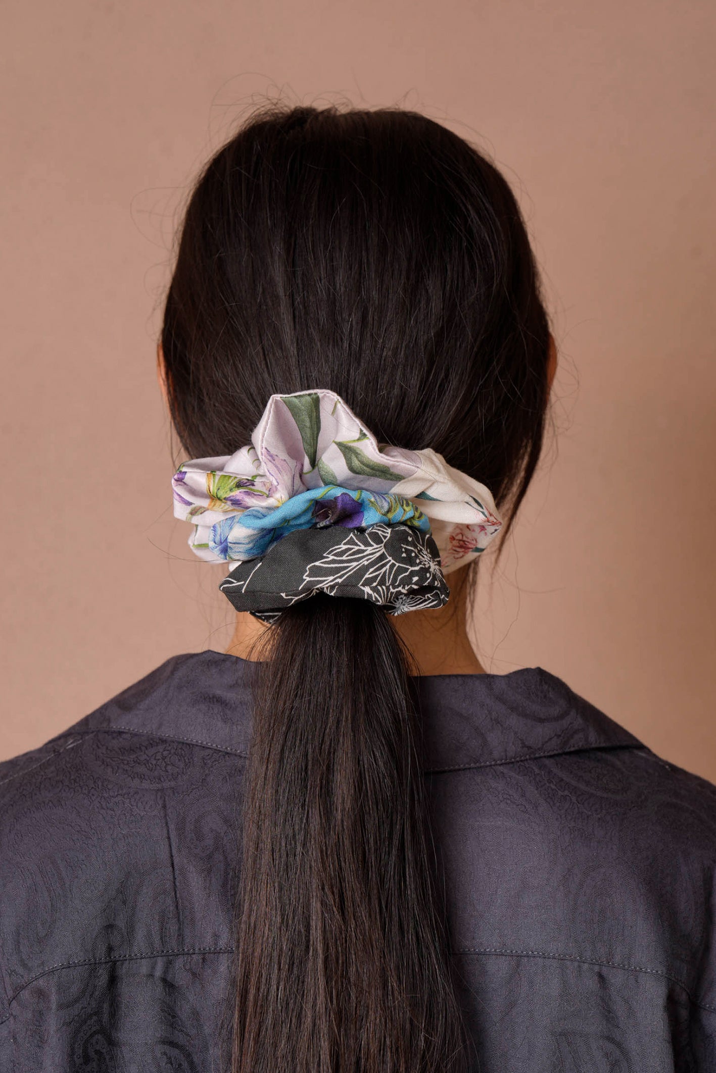 A woman with long, dark hair is seen from behind, wearing a mix of colorful scrunchies in floral prints featuring lilac, cyan, and black-and-white hues. She pairs them with a dark, textured paisley shirt against a simple beige background, highlighting the playful charm of scrunchies.