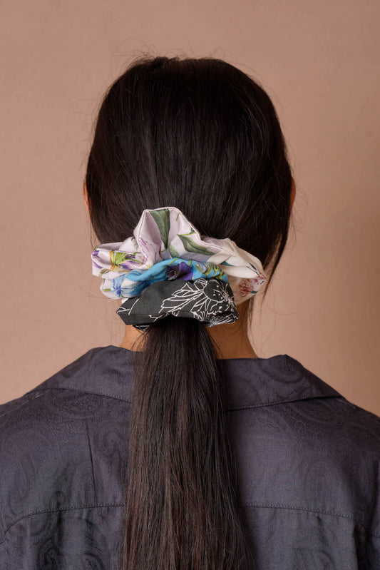 A woman with long, dark hair is seen from behind, wearing a mix of colorful scrunchies in floral prints featuring lilac, cyan, and black-and-white hues. She pairs them with a dark, textured paisley shirt against a simple beige background, highlighting the playful charm of scrunchies.
