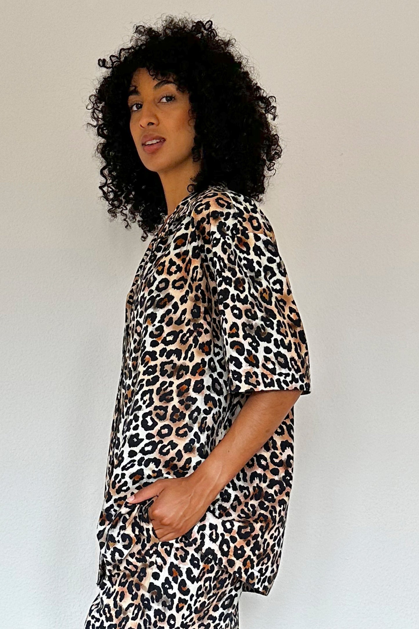 A person with curly hair stands against a plain background, wearing a matching leopard print loungewear set featuring a shirt with short sleeves and wide-legged pants. They are also wearing black sandals. The person looks towards the camera with a neutral expression. The floor is wooden.