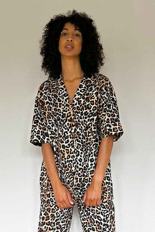 Woman with curly hair stands against a white wall on a wooden floor. She is wearing a matching leopard print set made from 100% organic cotton, featuring a short-sleeve top and wide-leg pants, along with black platform sandals. She looks directly at the camera with a relaxed posture.