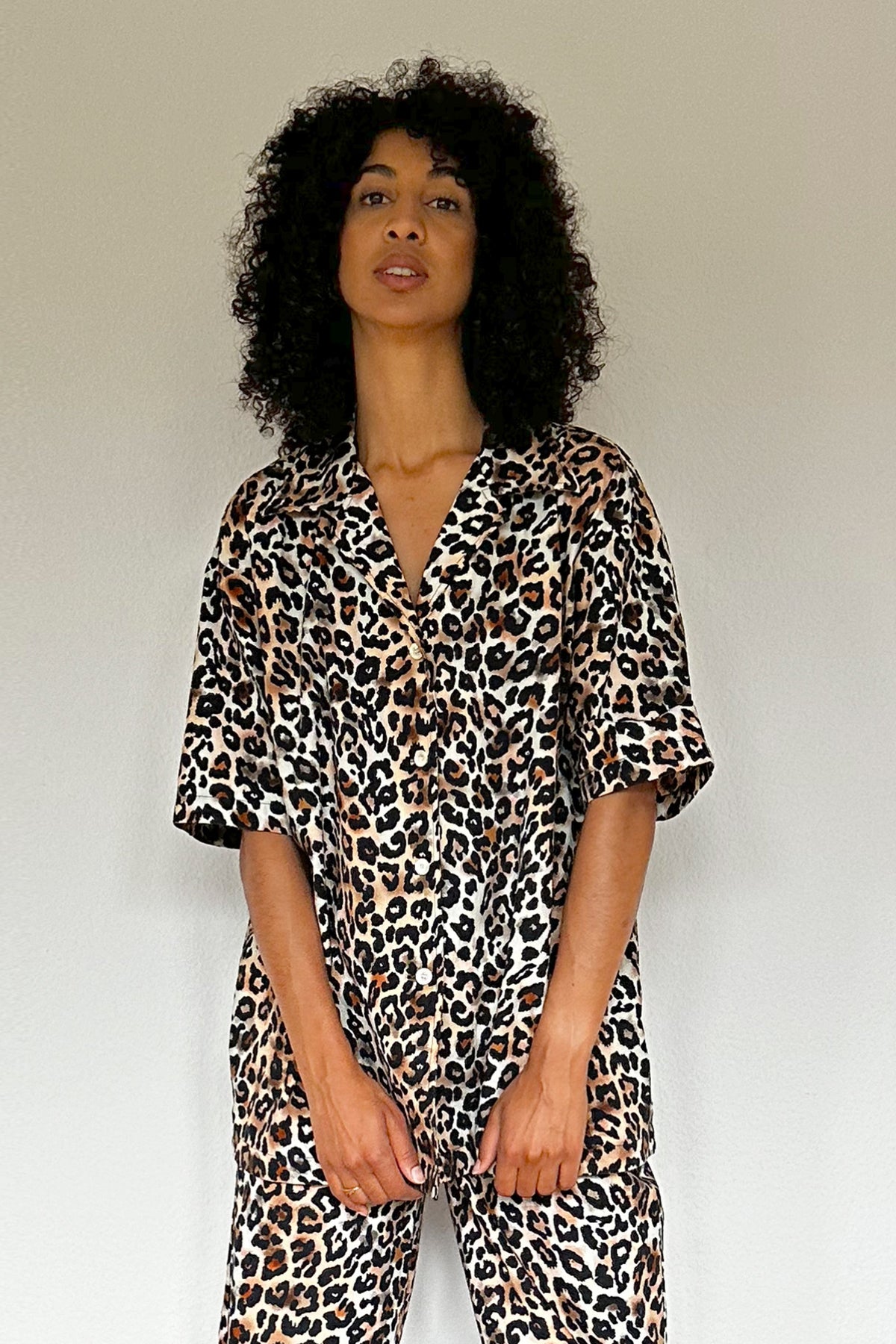 Woman with curly hair stands against a white wall on a wooden floor. She is wearing a matching leopard print set made from 100% organic cotton, featuring a short-sleeve top and wide-leg pants, along with black platform sandals. She looks directly at the camera with a relaxed posture.