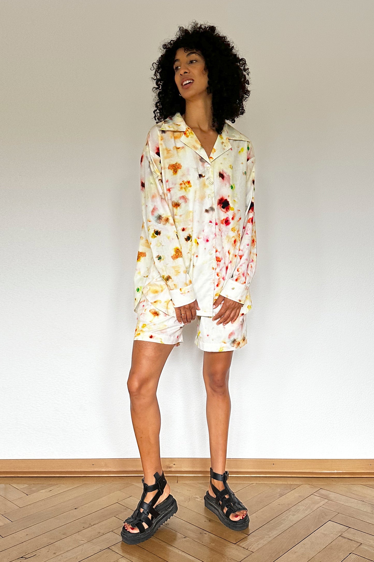 A person with curly hair is wearing a stylish loungewear set, consisting of a long-sleeve, collared blouse and matching shorts with a colorful abstract flower pattern. They are also wearing black strappy sandals. They stand against a plain white wall on a wooden floor, looking slightly to the side.