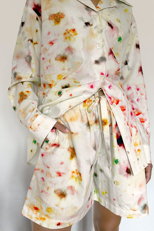A person is wearing a matching loungewear set of a long-sleeved button-up shirt and drawstring shorts. The outfit features a soft, off-white fabric with a colorful, abstract floral print in shades of yellow, orange, red, green, and black.