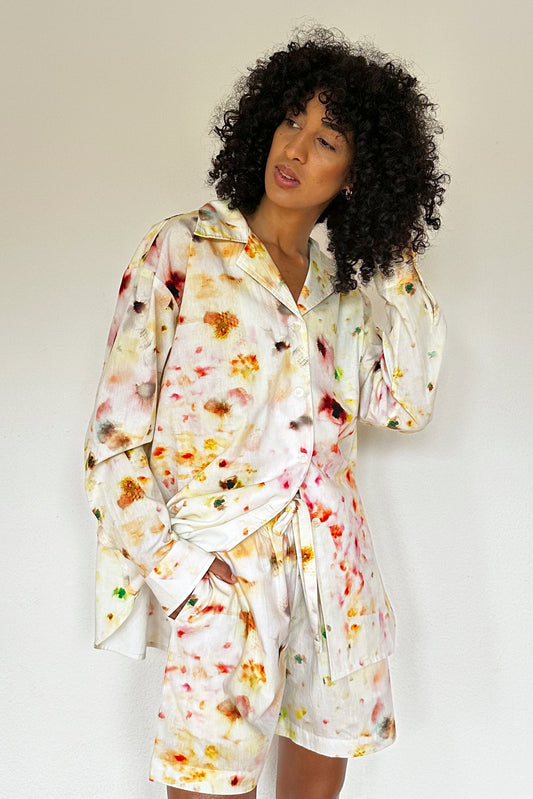 A person with curly hair dressed in a white loungewear set consisting of a shirt and matching shorts featuring a colorful, abstract flower pattern stands against a plain background. Their right hand is touching their hair while the left hand rests by their side.