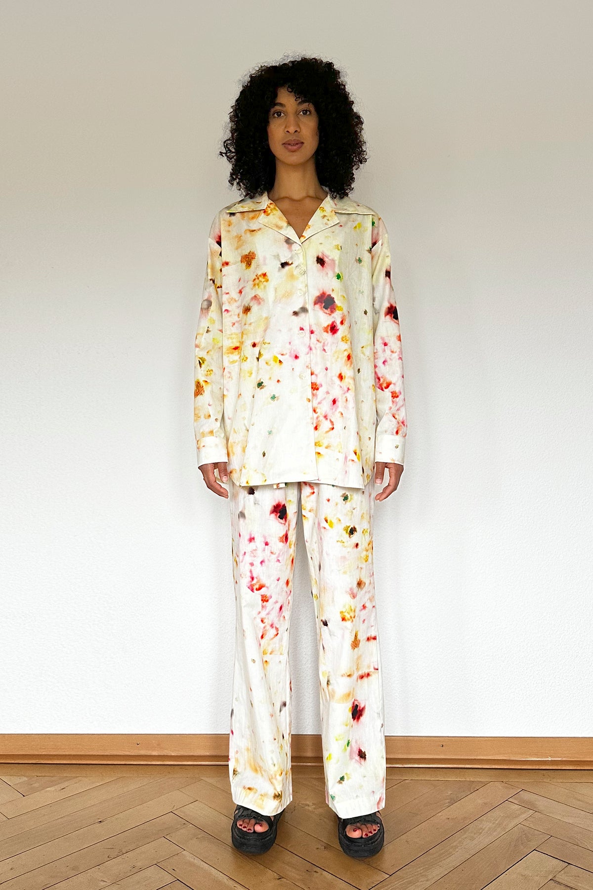A person stands against a white wall, wearing a matching loungewear set with a colorful blurry flower pattern featuring splashes of red, yellow, and green. The organic cotton set includes a long-sleeve, button-up shirt and loose-fitting pants. The person has curly hair and wears black sandals.