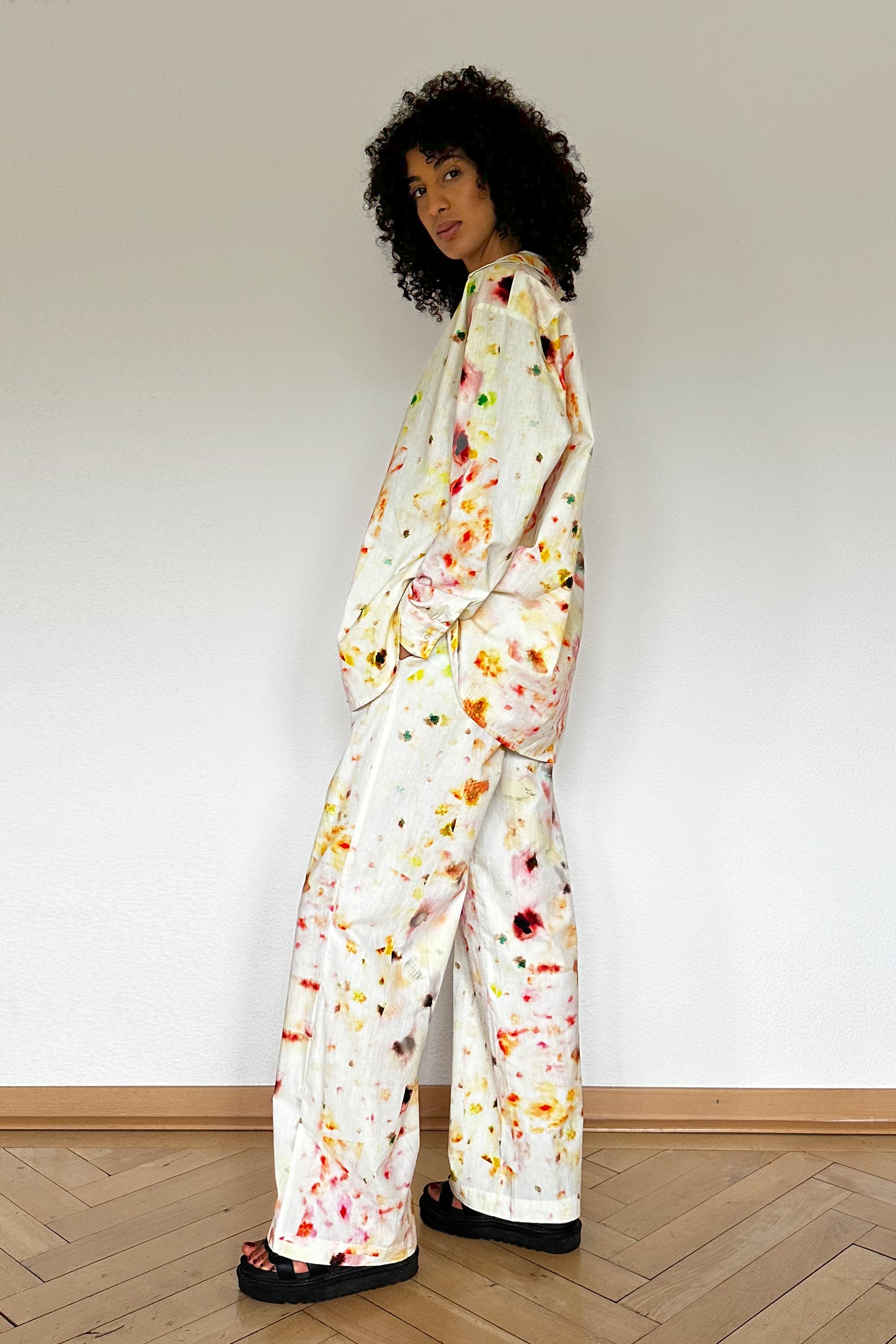 A person with curly hair stands against a plain white wall, wearing a loose-fitting loungewear set. The long-sleeved shirt and wide-legged pants feature a colorful paint splatter design. They have one hand in their pocket and are wearing black sandals, looking effortlessly stylish and comfortable.