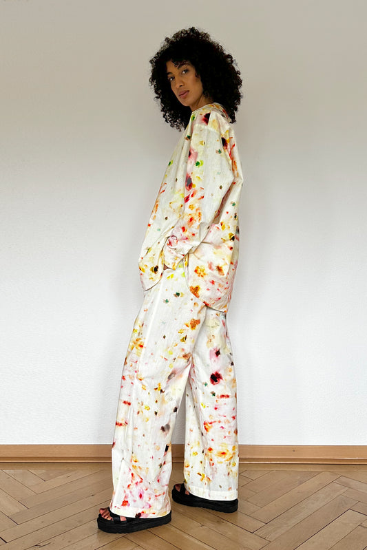 A person with curly hair stands against a plain white wall, wearing a loose-fitting loungewear set. The long-sleeved shirt and wide-legged pants feature a colorful paint splatter design. They have one hand in their pocket and are wearing black sandals, looking effortlessly stylish and comfortable.
