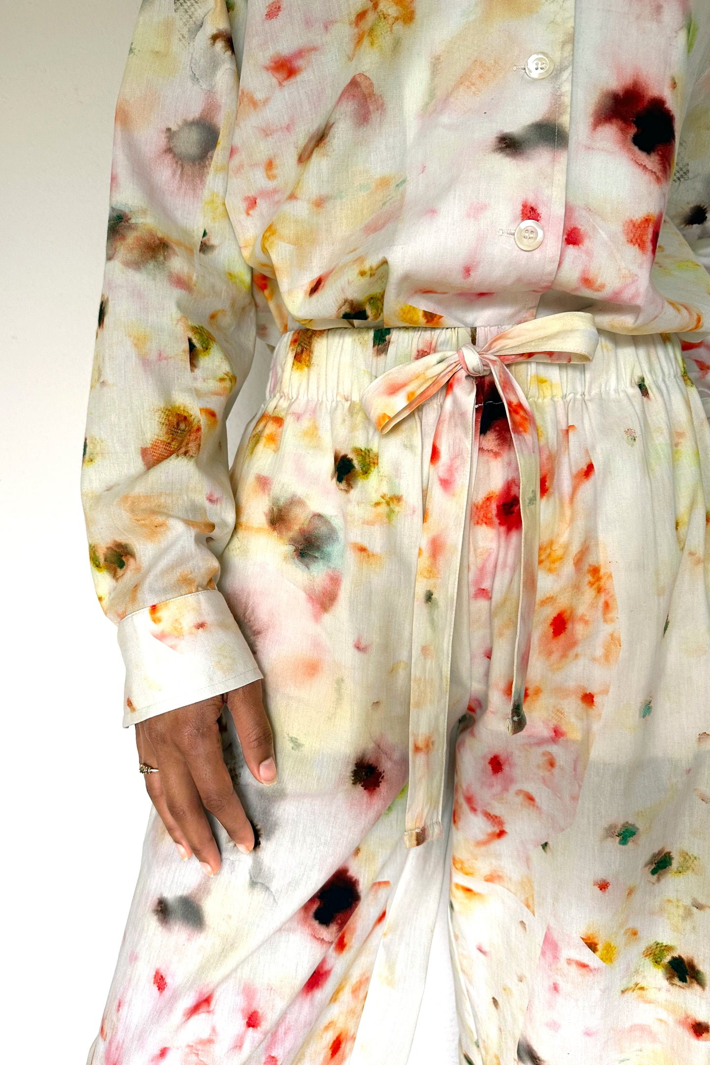 A person is wearing a matching loungewear set with a white base and an abstract, multicolored, blurry flower pattern. Their left hand hangs by their side, showing a ring on their finger. The background is a plain, neutral color.