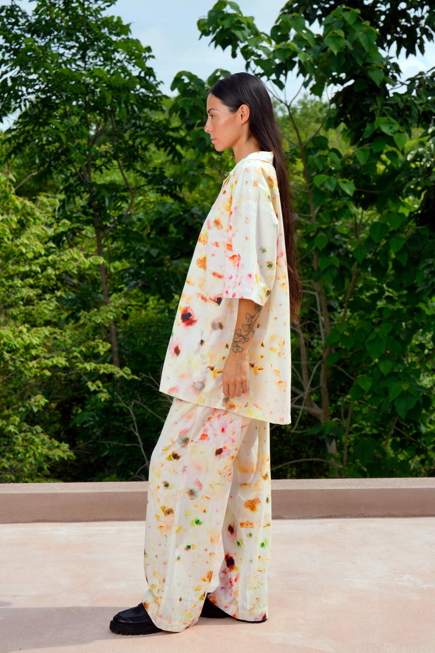 Side profile of a model wearing the pajama set in a blurry flower print made from organic cotton, standing outdoors against a backdrop of lush greenery.