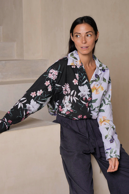 A woman leans casually on a beige stone ledge, wearing a bold, contrasting tropical flower print PJ-style blouse made from organic cotton. The flowing jacquard paisley pants add texture, blending comfort and sophistication in this modern loungewear ensemble.