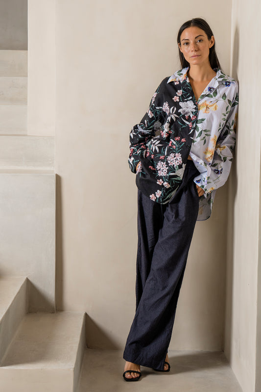 A model stands beside a concrete staircase, wearing a loungewear PJ set featuring a tropical floral print split into two colors—black on one side and lilac on the other. The relaxed-fit shirt pairs effortlessly with deep navy jacquard paisley pants, creating a chic yet comfortable look. Styled with black heels, this ensemble perfectly embodies loungewear designed for both home and beyond.