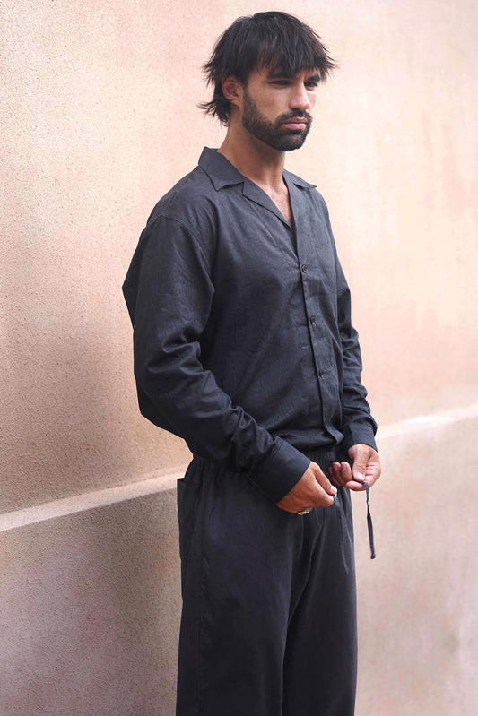 A man stands against a wall in a black loungewear set for men, featuring a matching shirt and pants in a jacquard paisley fabric. He gazes contemplatively to the right while adjusting the drawstring of his pants.