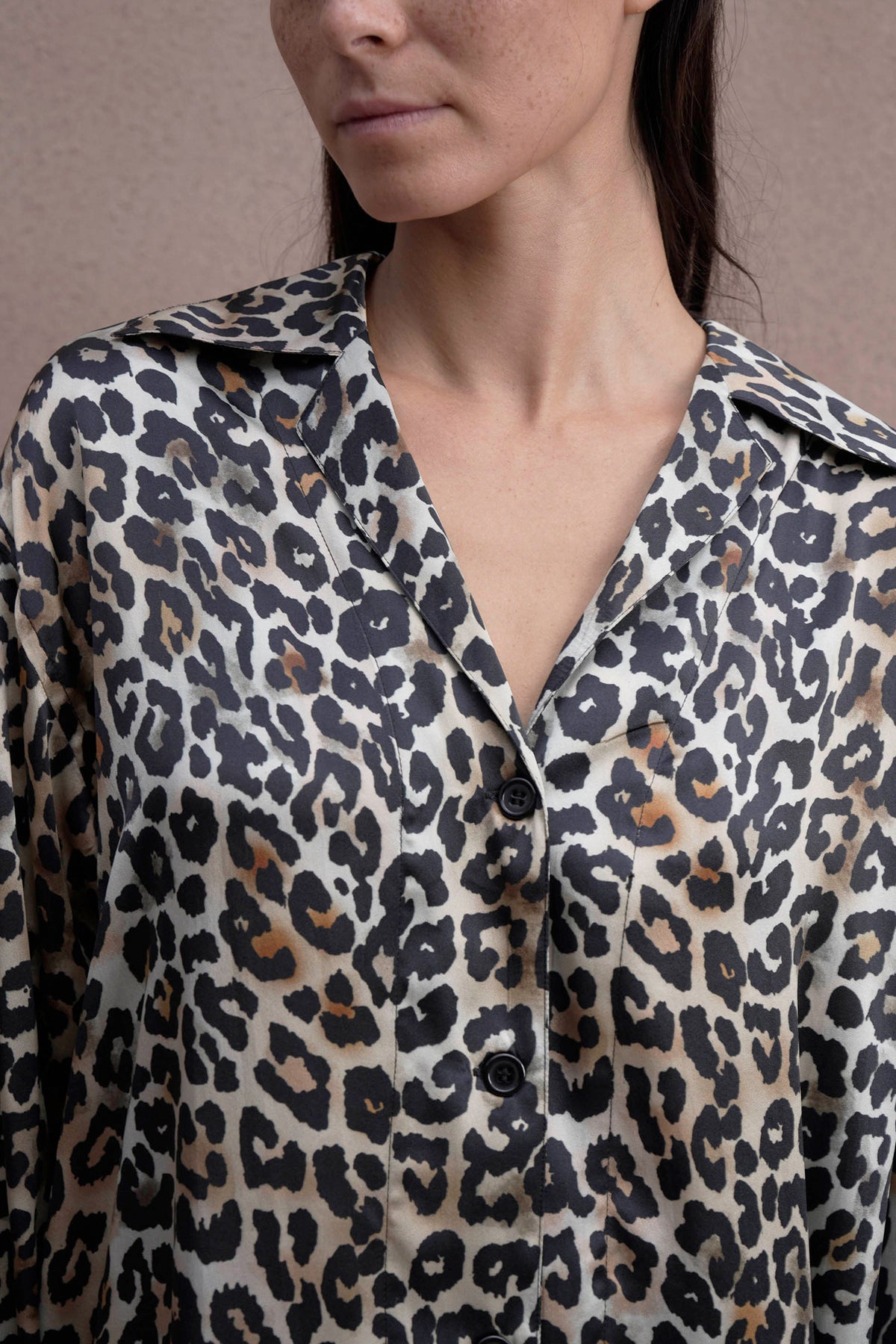 A person wearing a button-up shirt in luxurious silk satin, showcasing a classic leopard print pattern. The shirt features a collar and comes in elegant shades of black, white, and brown. The background is a neutral beige color.