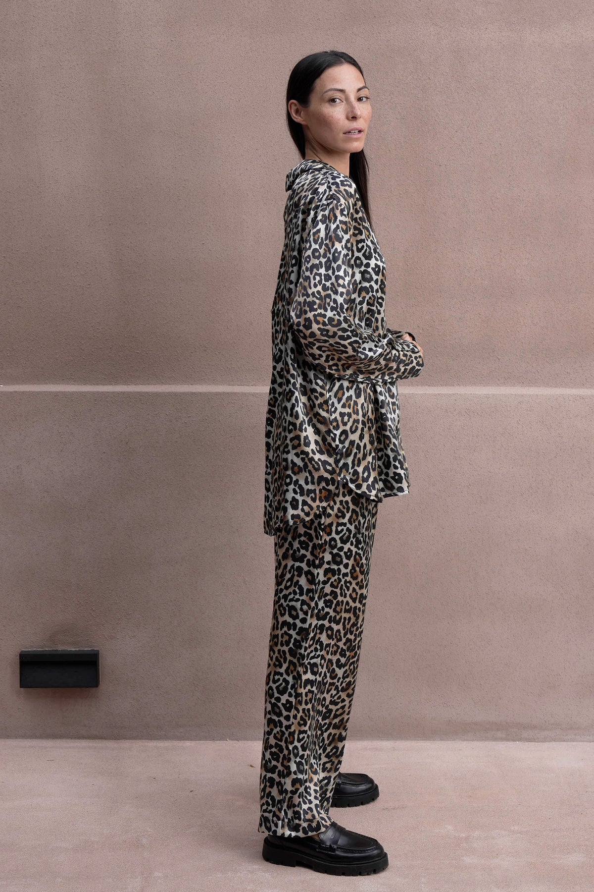 A woman with long, dark hair and a confident expression wears a leopard print loungewear set with a matching shirt and pants. The silk satin fabric drapes elegantly, creating a relaxed yet sophisticated look. She stands against a neutral-toned wall, pairing the outfit with chunky black loafers for a modern edge.