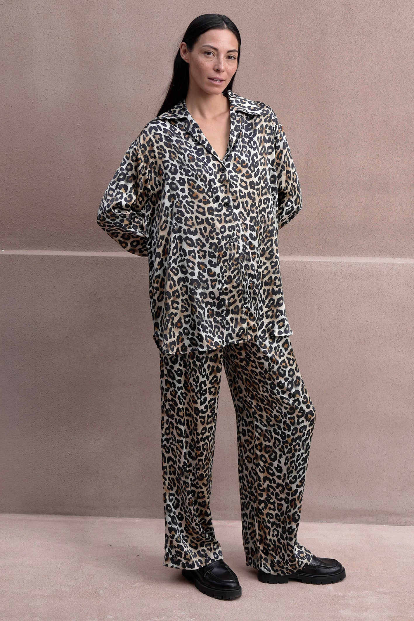 A person stands against a beige wall in a silk satin leopard print pajama set with a button-up shirt and matching pants. Their long black hair flows down, and they complete the look with black shoes.