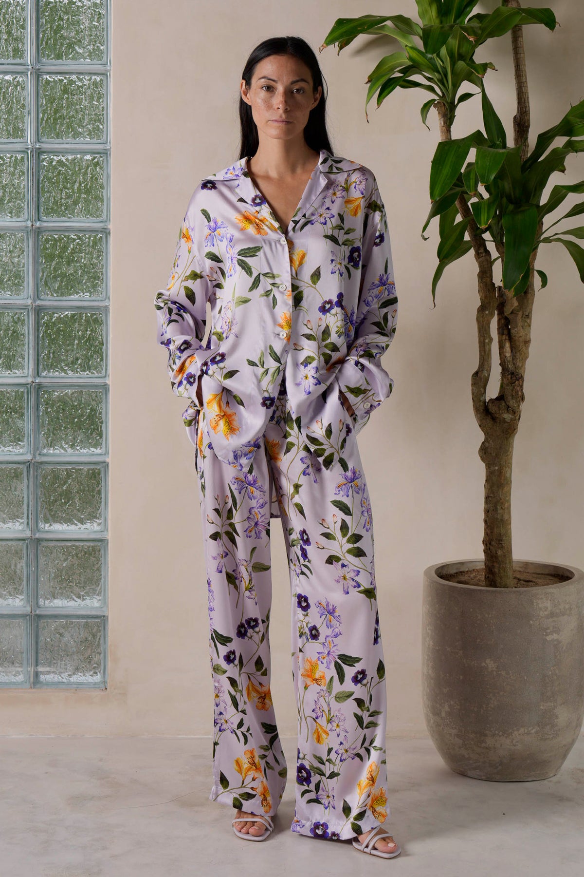 A woman stands indoors wearing a silk loungewear set with a lilac meadow flower print. The matching shirt and pants have a soft lilac background adorned with colorful floral patterns. She pairs the look with strappy sandals, standing near a potted plant and a glass block wall.