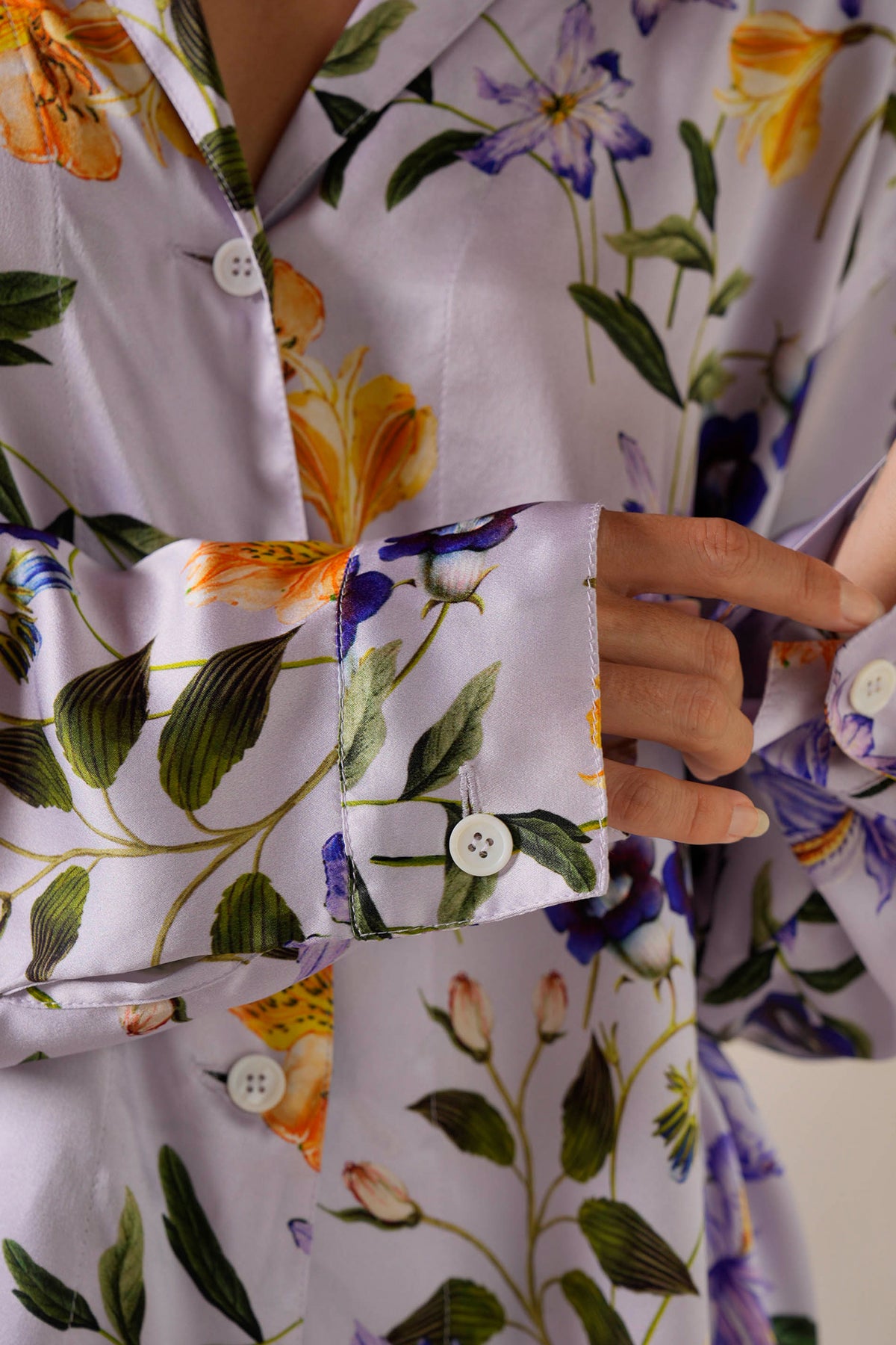 Close-up of a woman wearing a long-sleeved satin shirt adorned with a lilac meadow flower print. The design features orange and purple flowers with green leaves, reminiscent of luxurious silk loungewear. The individual is buttoning a cuff with white buttons.