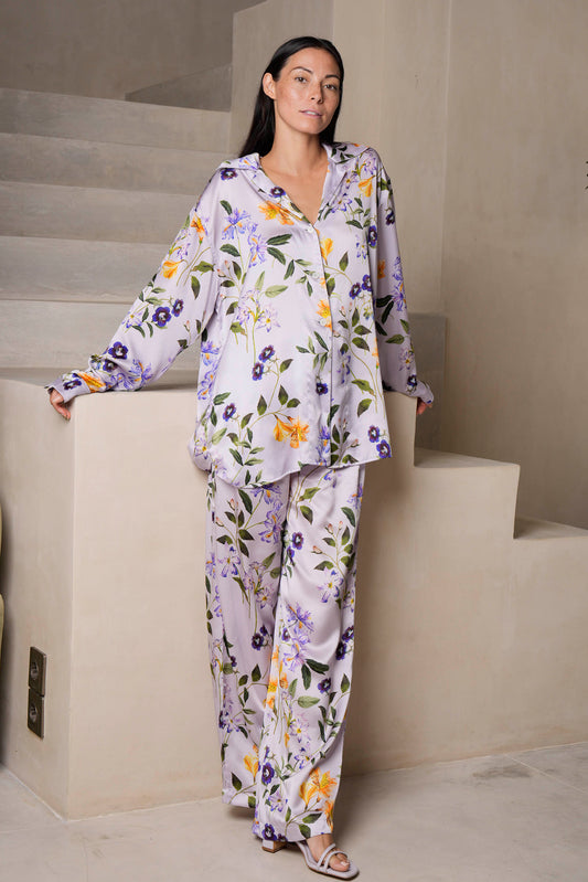 A woman with long dark hair leans on light-colored stairs, wearing a silk loungewear set featuring a lilac meadow flower print. The loose-fitting shirt and pants complement her strappy sandals as she casually rests against the wall with a neutral expression.