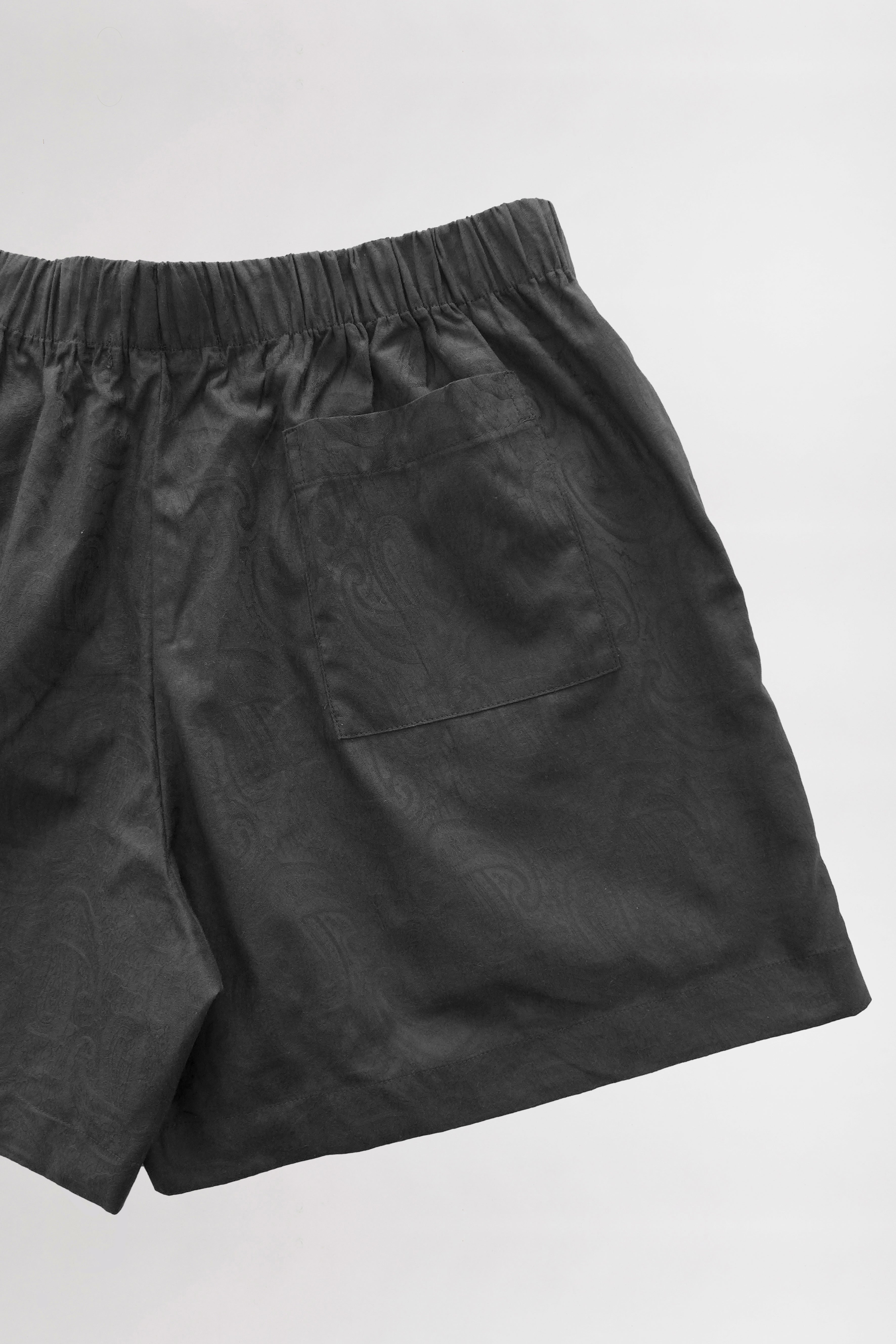 A pair of black, unisex shorts with an elastic waistband and a single back pocket. The shorts appear to be made of lightweight Cotton Jacquard Paisley fabric. The background is a clean, plain white surface.