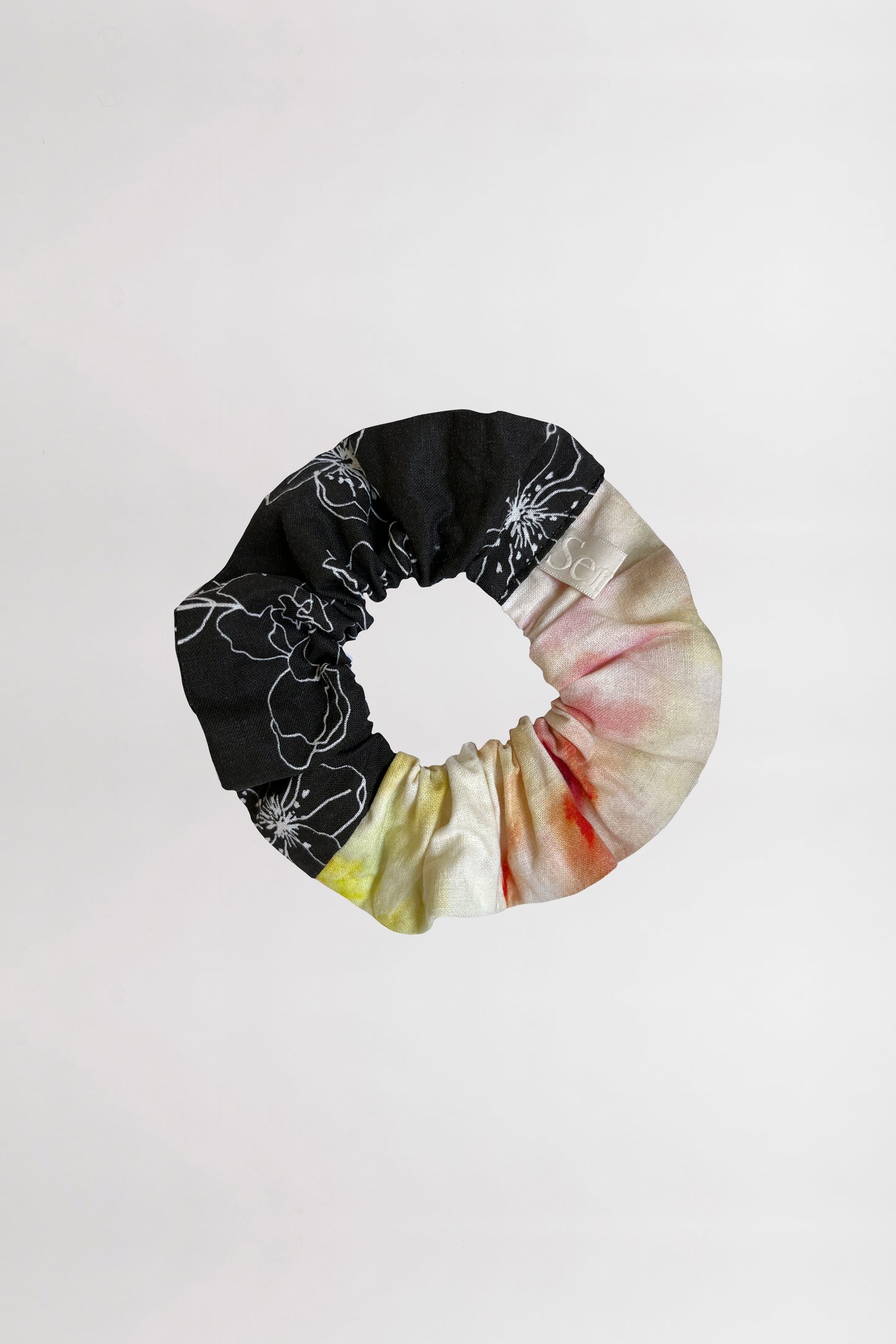 Front view of a scrunchie featuring two unique patterns – one with white linear flowers on a black background, and the other with a colorful abstract design – crafted from leftover fabrics, set against a neutral backdrop.