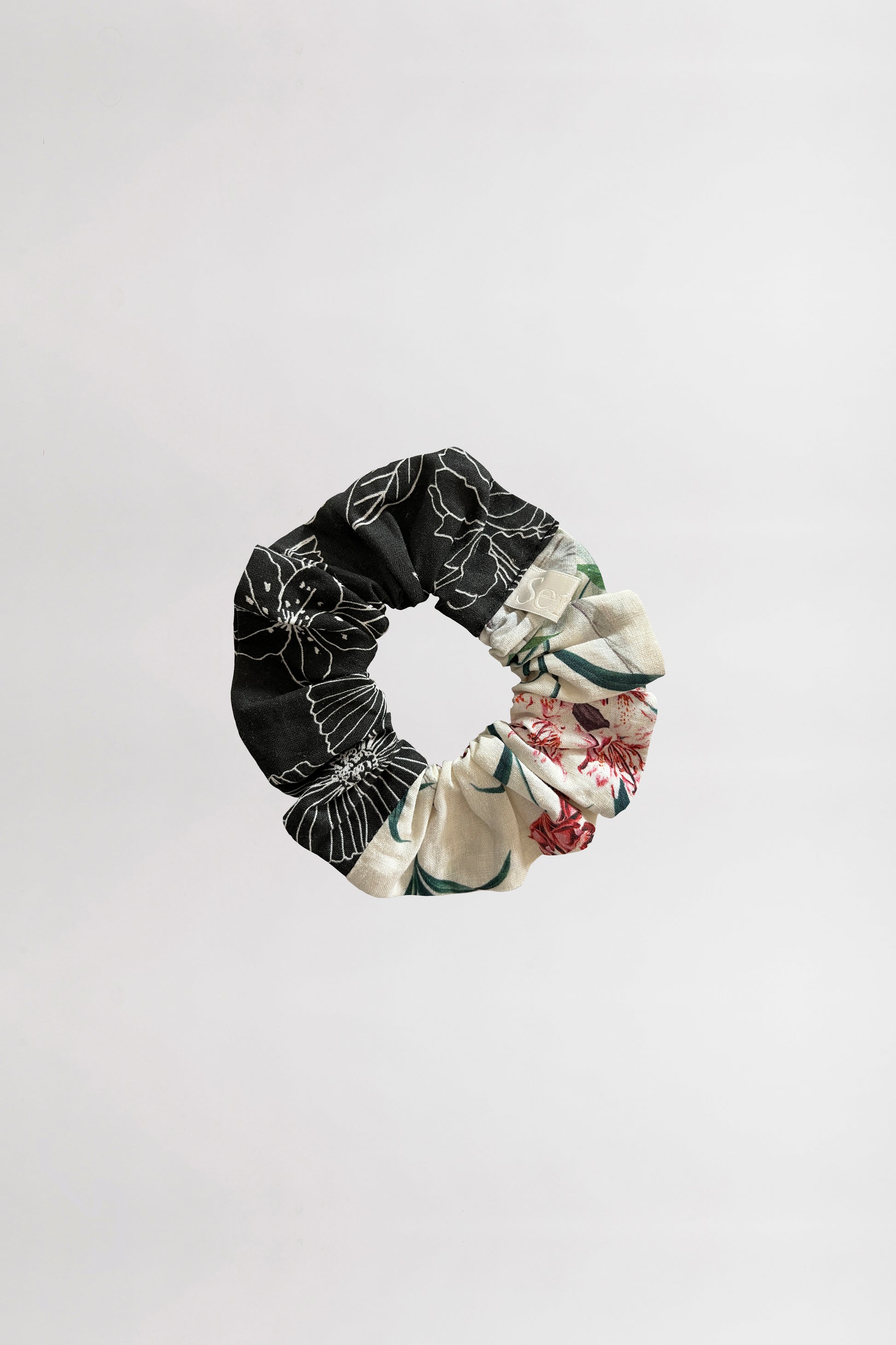 Front view of a scrunchie featuring two unique floral designs, one black with white linear flowers and one with a cream background and tropical flowers, crafted from leftover fabrics against a neutral background.