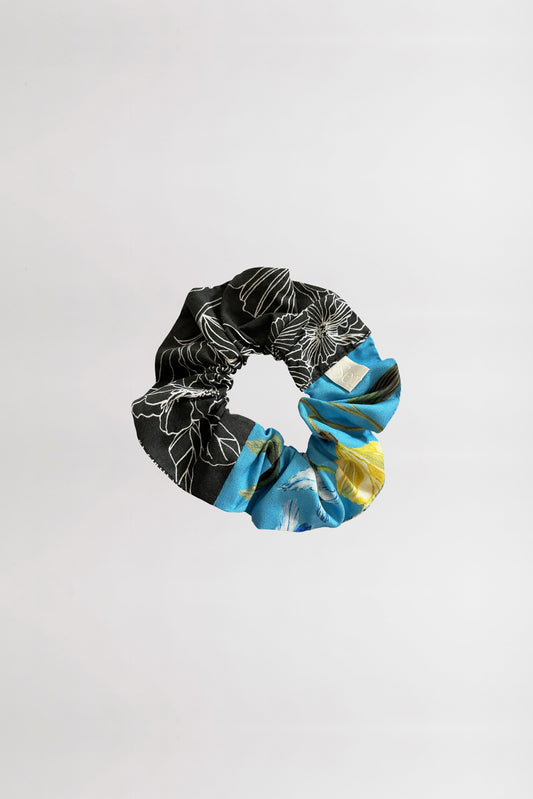 Front view of a scrunchie showcasing two floral patterns: one with a black background and white linear flowers, the other with a sky blue background and tropical blooms, made from leftover fabrics on a neutral backdrop.