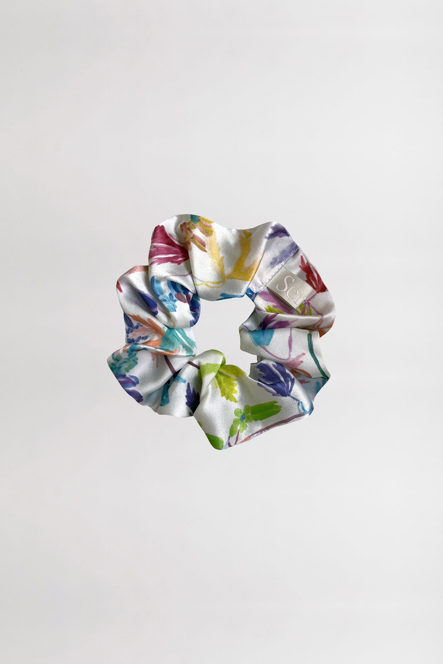 A silk satin scrunchie with a vibrant flower print, featuring an array of colors such as green, blue, purple, and pink, is displayed against a plain white background. Made from leftover fabric, the scrunchie is crumpled in a circular shape, beautifully showcasing its colorful design.