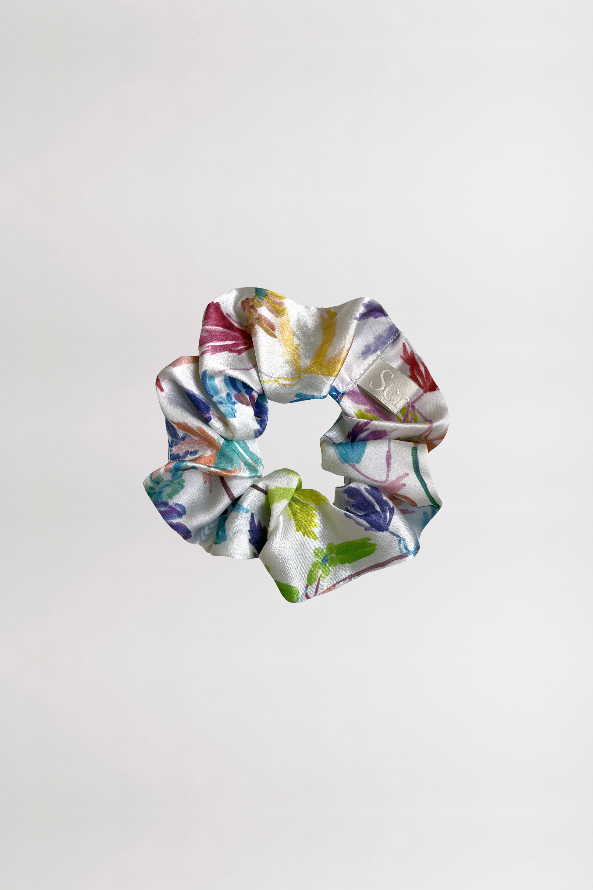 A silk satin scrunchie with a vibrant flower print, featuring an array of colors such as green, blue, purple, and pink, is displayed against a plain white background. Made from leftover fabric, the scrunchie is crumpled in a circular shape, beautifully showcasing its colorful design.