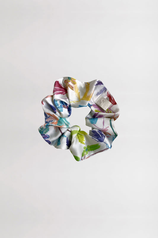 A silk satin scrunchie with a vibrant flower print, featuring an array of colors such as green, blue, purple, and pink, is displayed against a plain white background. Made from leftover fabric, the scrunchie is crumpled in a circular shape, beautifully showcasing its colorful design.
