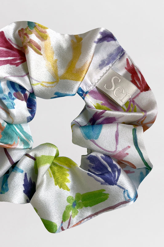 A silk satin scrunchie with a colorful floral and leaf pattern on a white background. The scrunchie, made from leftover fabric, has a small sewn-on label with a the Serlet logo. The fabric appears soft and glossy, creating a slightly crinkled texture.