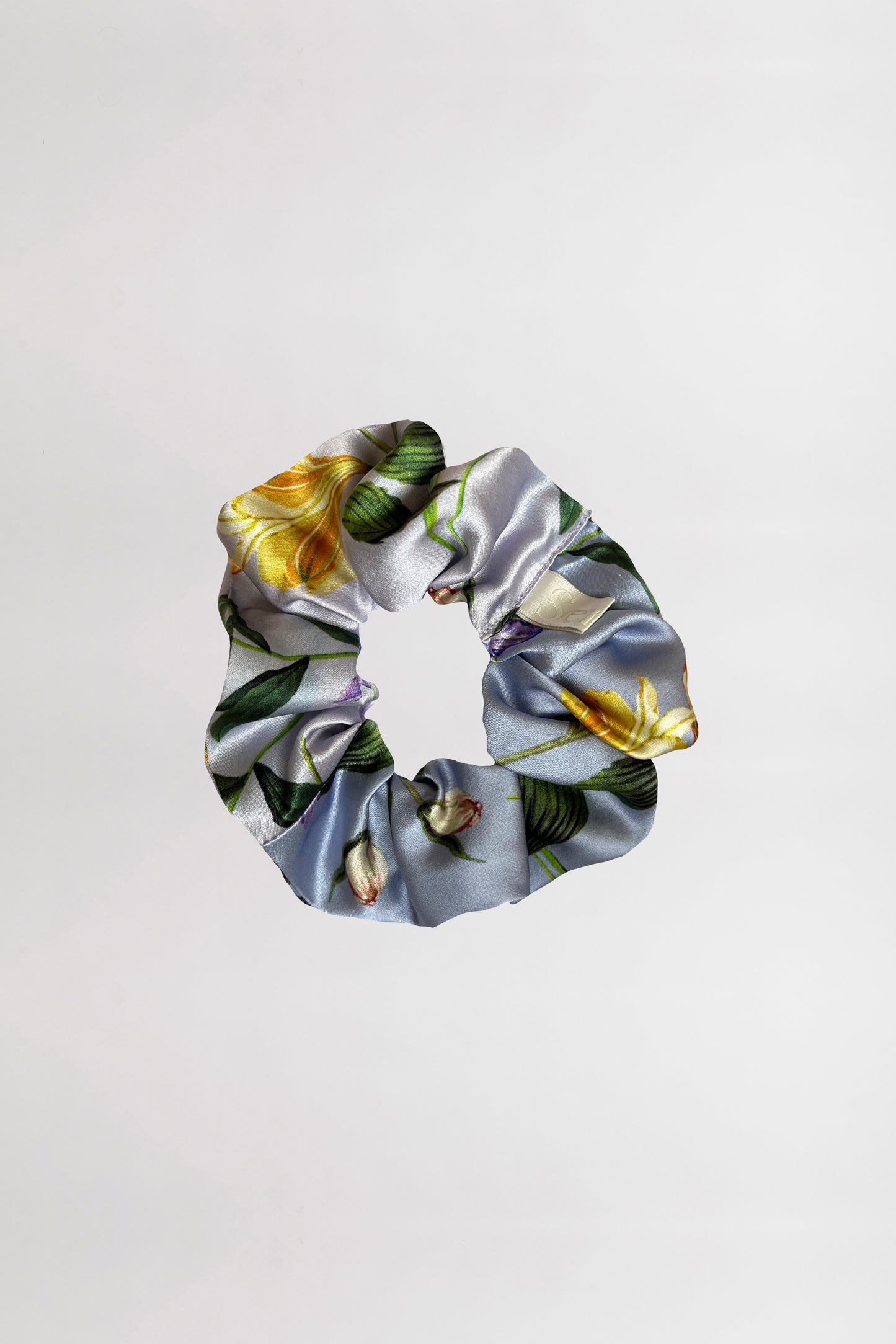 A silk satin scrunchie made of shiny silk satin fabric with a floral pattern featuring green leaves and yellow flowers, set against a light gray background. This elegant scrunchie, created from left over fabric, is centered against a plain white backdrop.