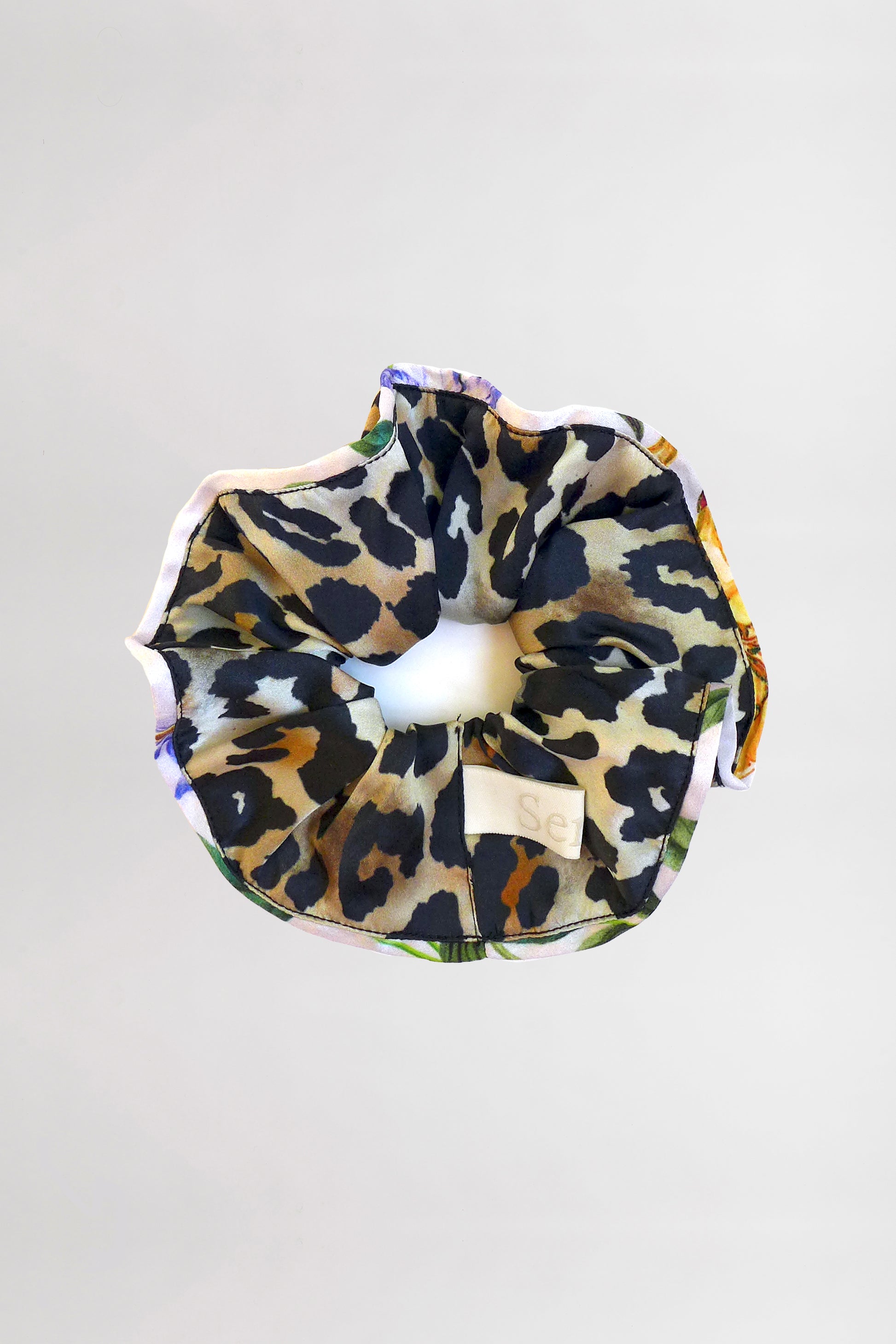 A Piped-Edge Silk Satin Scrunchie with a leopard print design lies against a plain light gray background. The fabric features black, tan, and beige spots and has a slight sheen. A small white tag with text is attached on one side.