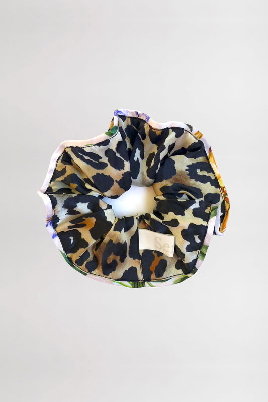 A Piped-Edge Silk Satin Scrunchie with a leopard print design lies against a plain light gray background. The fabric features black, tan, and beige spots and has a slight sheen. A small white tag with text is attached on one side.
