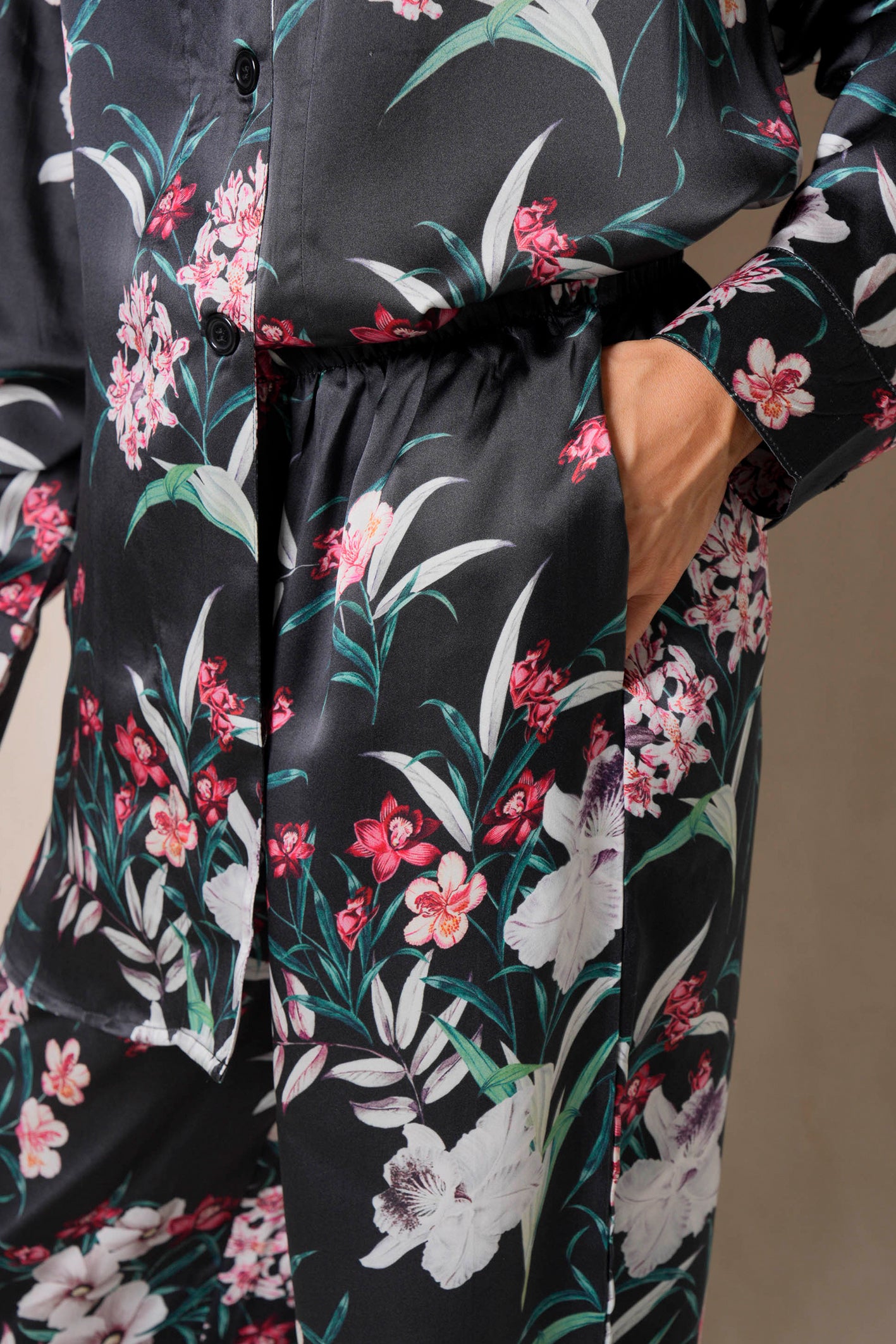 A person is dressed in a black silk loungewear set featuring a vibrant tropical flower print with red, pink, and white blooms alongside green leaves. The ensemble includes a button-up shirt and pants, with the person's hand casually resting in one of the pockets.