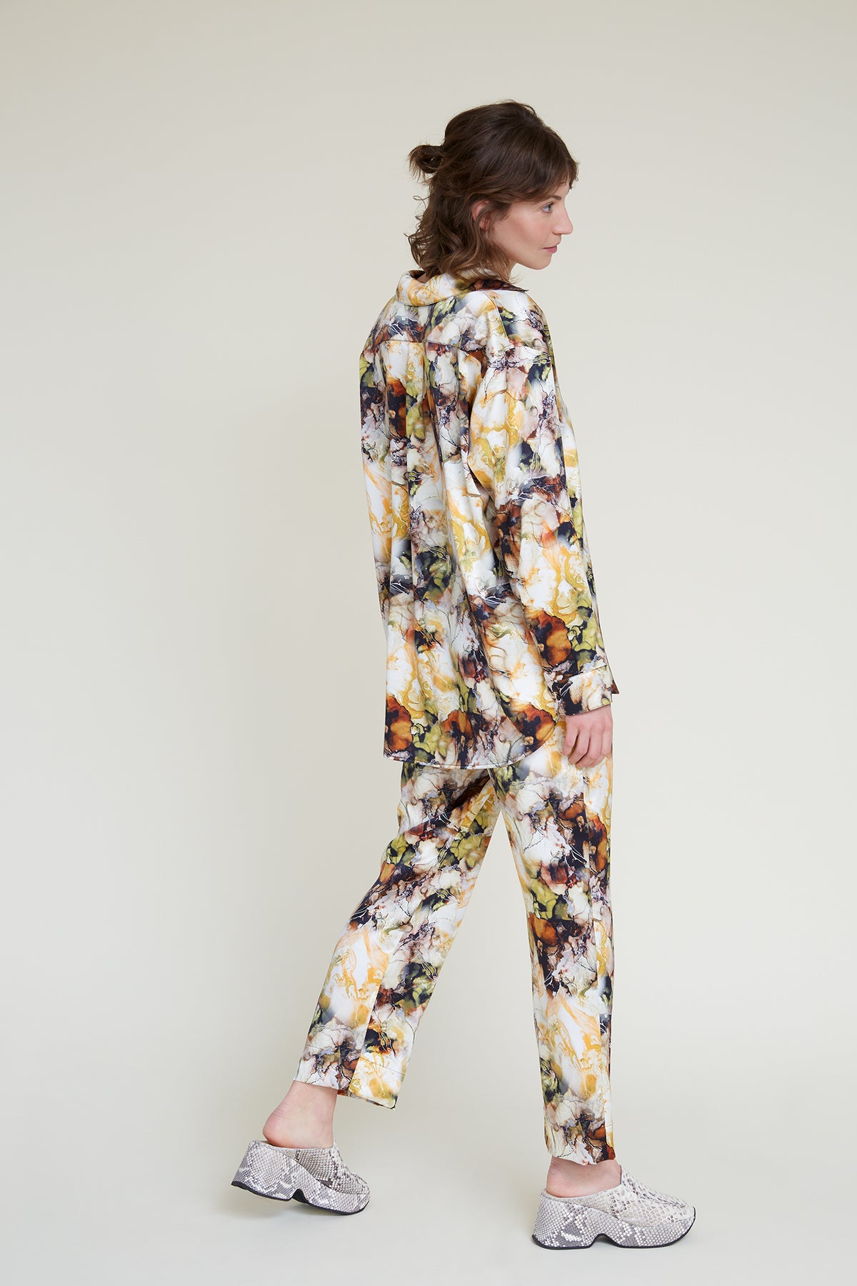 A person with shoulder-length hair is seen from the side, wearing a matching shirt and pants in a colorful, abstract pattern. The loungewear set is made from luxurious silk satin fabric that mimics the look of marbled prints. They are also wearing light-colored, textured shoes with a thick sole. The background is plain and light-colored.