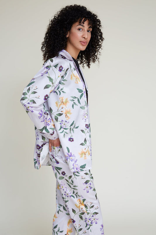 A person with curly hair is posing in a floral-patterned loungewear set consisting of a loose long-sleeved top and matching pants. The background is plain and light-colored. The person has one hand on their hip and is turned slightly to the side, looking toward the camera.