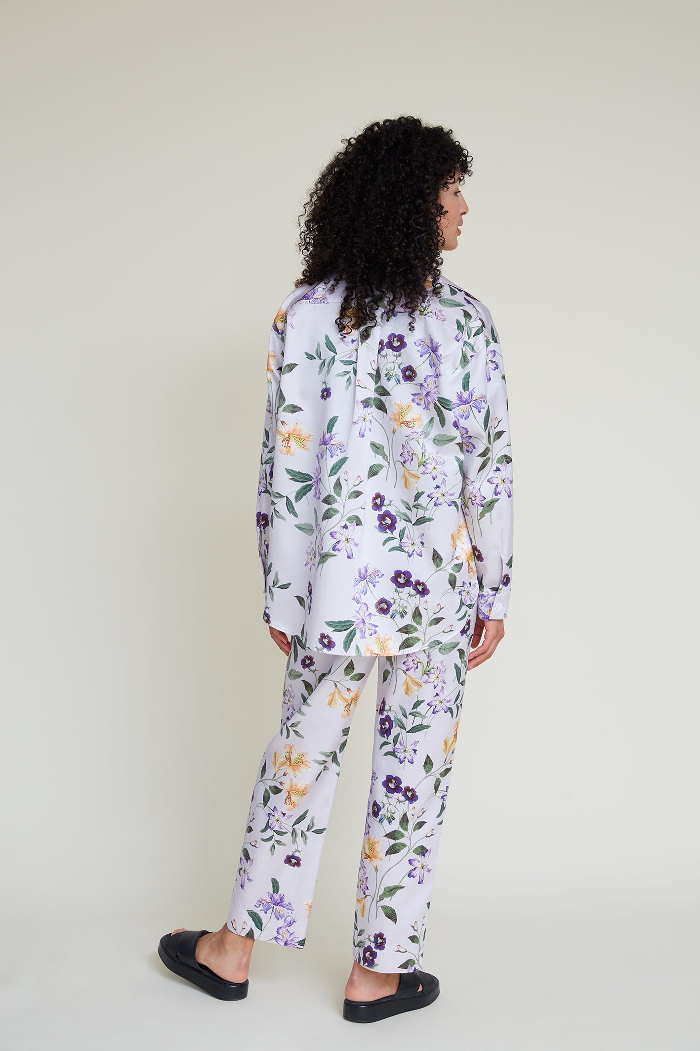 A person with curly hair stands facing away, dressed in a lilac loungewear set featuring a floral pattern in shades of purple, yellow, and green. They wear black slip-on shoes against a plain background.