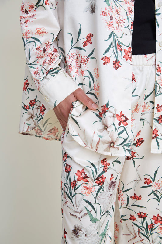 Close-up of a person wearing a floral-patterned loungewear set. The hand is tucked into the side pocket of the loose-fitting pants. The pajama fabric is predominantly off-white with a detailed print of red, pink, and brown flowers and green leaves.