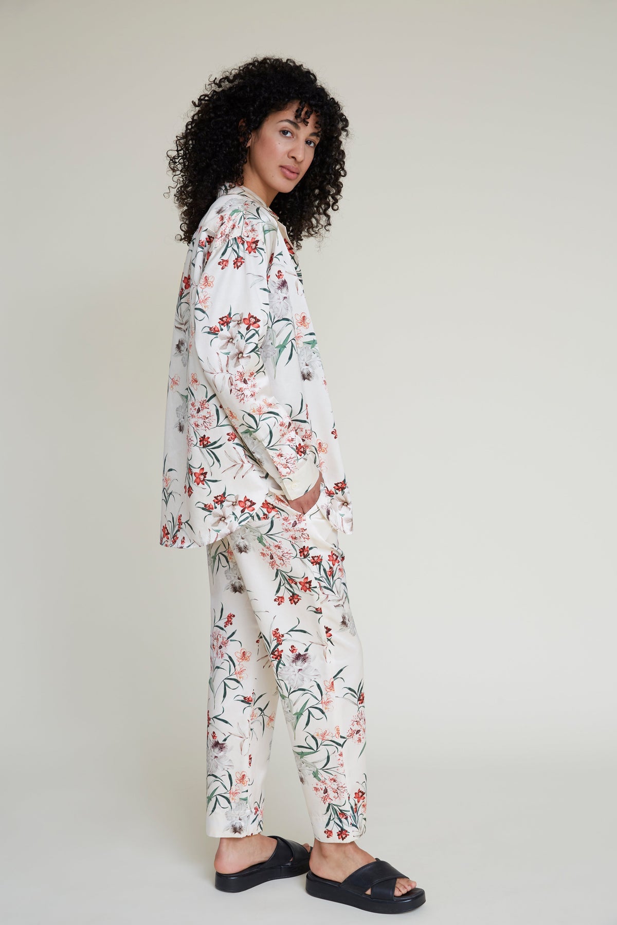 A person with curly hair stands against a plain background, wearing a matching tropical floral-patterned loungewear set consisting of a long-sleeved shirt and pants. They are looking over their shoulder with hands in pockets and wearing black slide sandals.