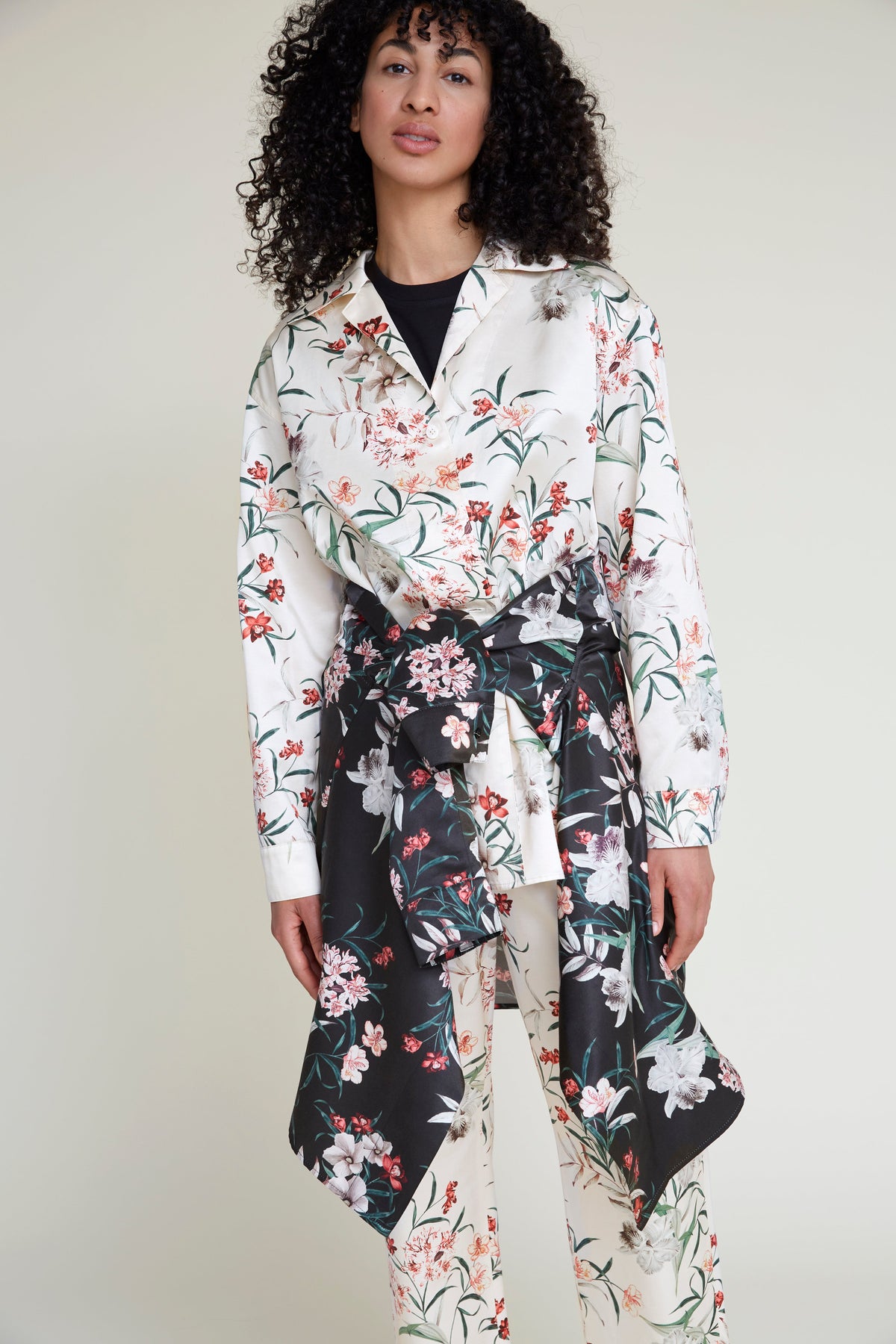 A woman with curly hair models a fashionable loungewear outfit. She wears a tropical flower print cream blouse and matching pants. A shirt with a black floral print is tied around her waist, accentuating the elegant yet stylish pajama set. The background is a plain, light color, highlighting her ensemble's sophisticated charm.