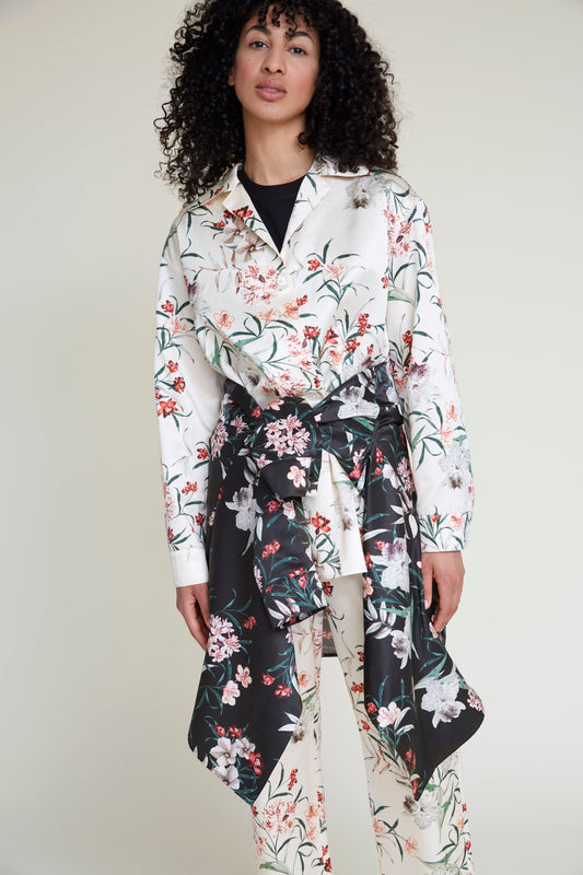 A woman with curly hair models a fashionable loungewear outfit. She wears a tropical flower print cream blouse and matching pants. A shirt with a black floral print is tied around her waist, accentuating the elegant yet stylish pajama set. The background is a plain, light color, highlighting her ensemble's sophisticated charm.