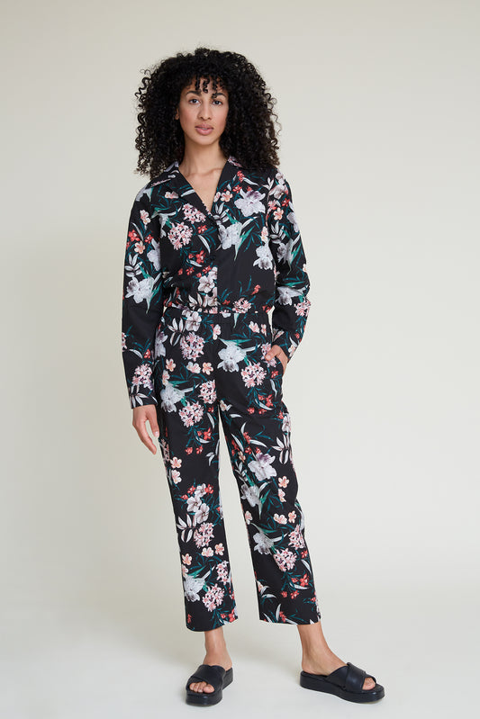 A person with curly hair is standing against a plain backdrop wearing a black loungewear set adorned with colorful tropical floral patterns. They have one hand in their pocket, wear black slide sandals, and exude the comfort of chic loungewear.