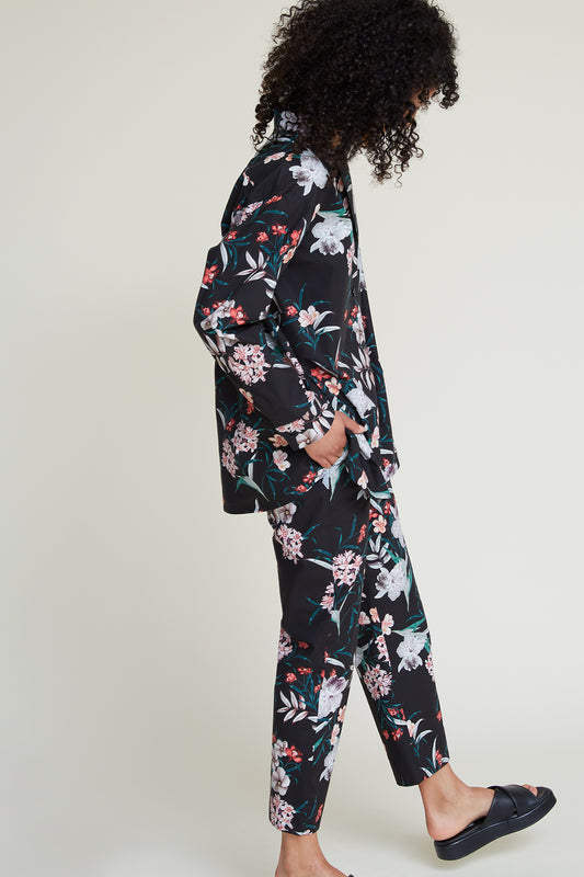 A person with curly hair is shown in profile, wearing a black loungewear set with a vibrant tropical floral pattern. They are standing against a plain white background and appear to be looking down. The outfit consists of a long-sleeve shirt and matching pants.