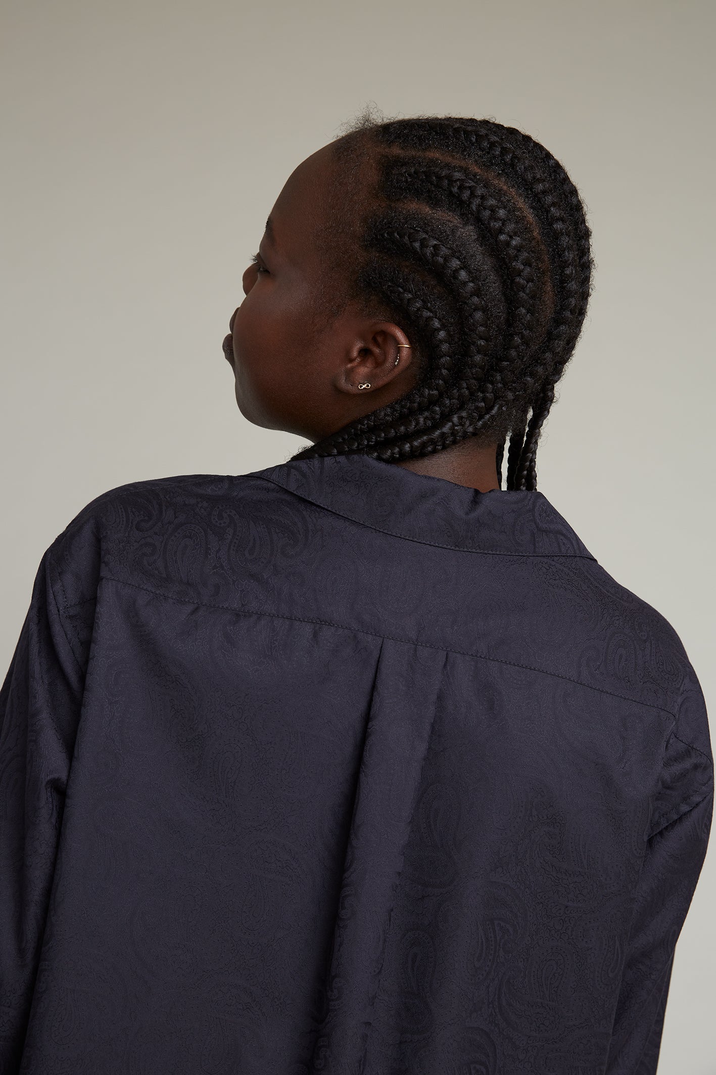A person with neatly braided hair is turned away, wearing a dark, textured jacquard paisley pajama shirt. The background is plain and neutral, allowing focus on the detailed braiding and fabric patterns.