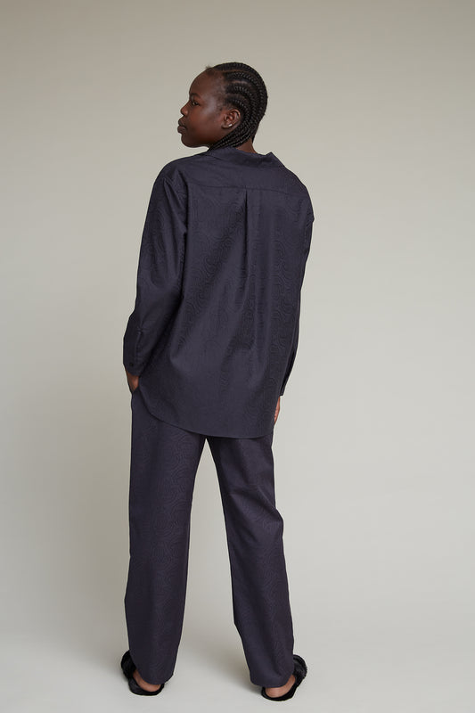 A person with braided hair is standing with their back to the camera, wearing a dark, paisley jacquard patterned, long-sleeved oversized shirt and matching pants. This stylish loungewear set contrasts softly against the plain, light-colored background.