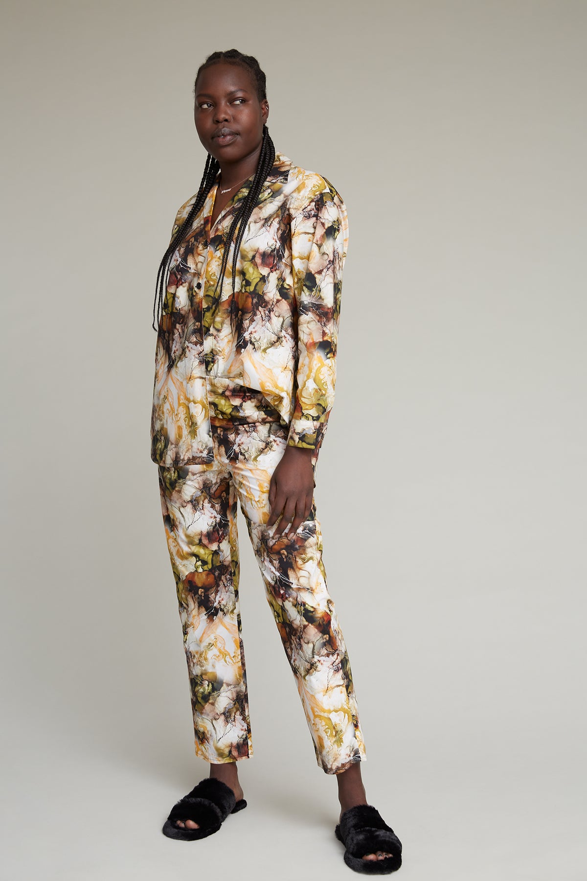 A person with long braided hair stands against a plain background wearing a colorful marble printed loungewear set. They are also sporting black, furry slippers. The individual has a confident expression and a relaxed posture.