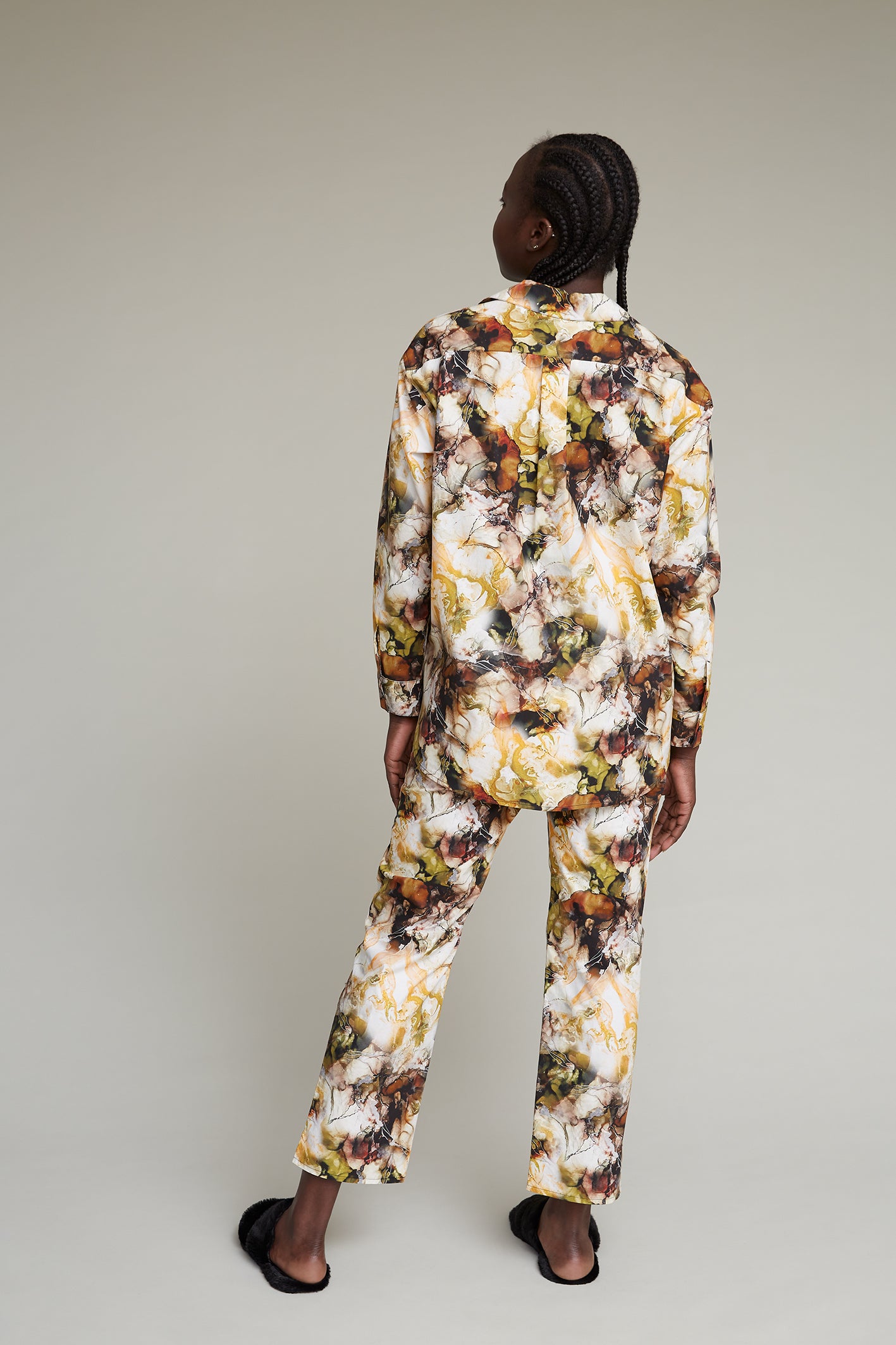 A person with braided hair stands facing away from the camera, wearing matching pants and a long-sleeved shirt with an abstract, multicolored marble print. The background is a plain, light gray. The person is also wearing black sandals.