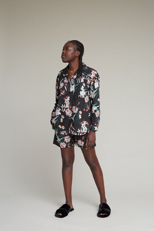 A person stands against a plain background, wearing a matching black tropical flower print loungewear set. The outfit is complemented by black slide-on sandals. The person has long, braided hair and appears relaxed with hands casually placed in the front pockets of their shorts.
