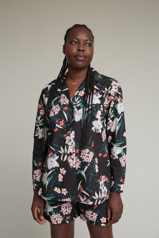 A person with long braids is wearing a matching floral loungewear set consisting of a button-up shirt and shorts. The clothing features a black background with colorful tropical flower prints. They have a neutral expression and are posed against a plain background.