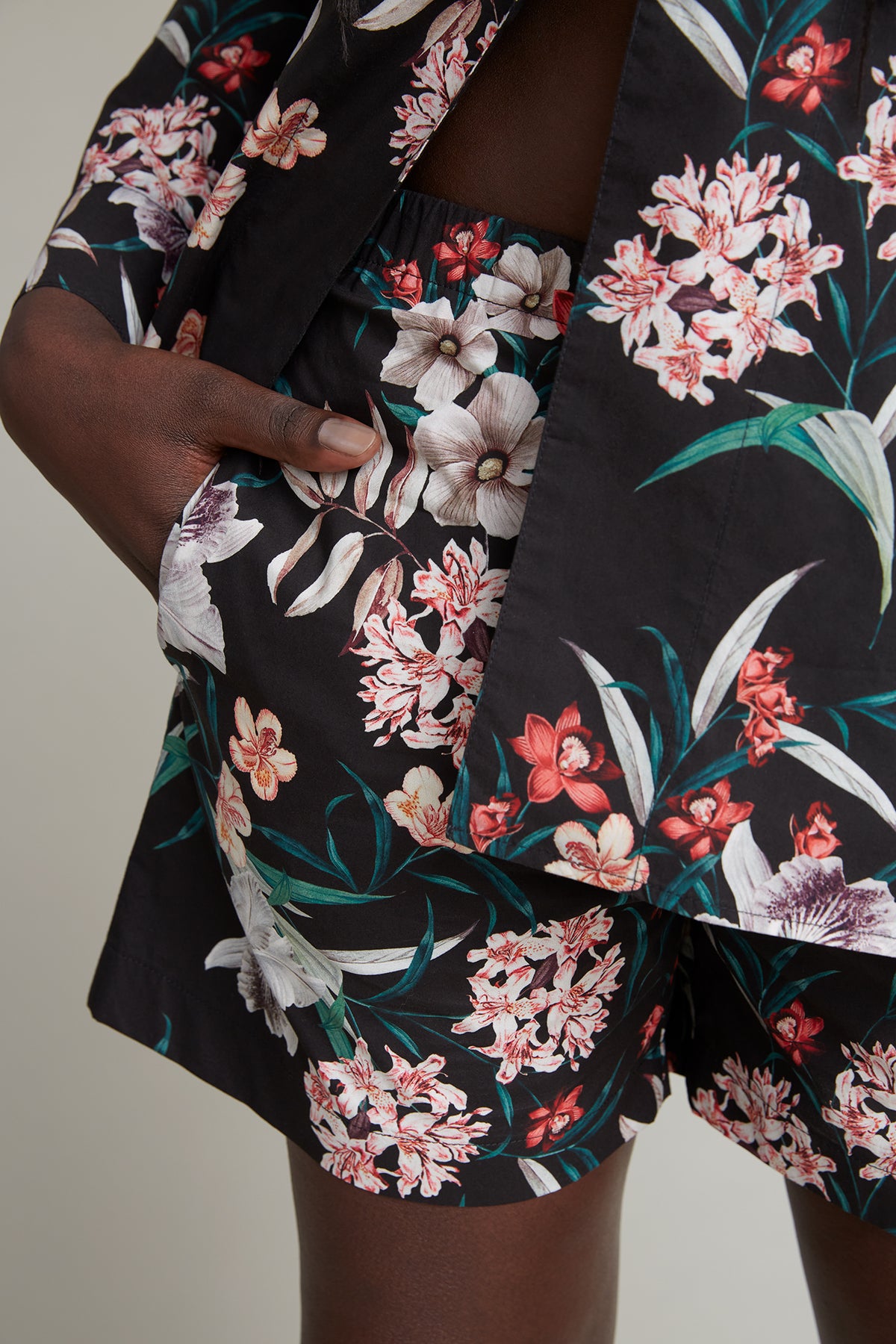 Close-up of a person wearing a black loungewear set adorned with a detailed tropical flower print featuring white, pink, and red blooms along with green leaves. With their hand partially in the pocket, the matching shirt and shorts create a cohesive and stylish look.